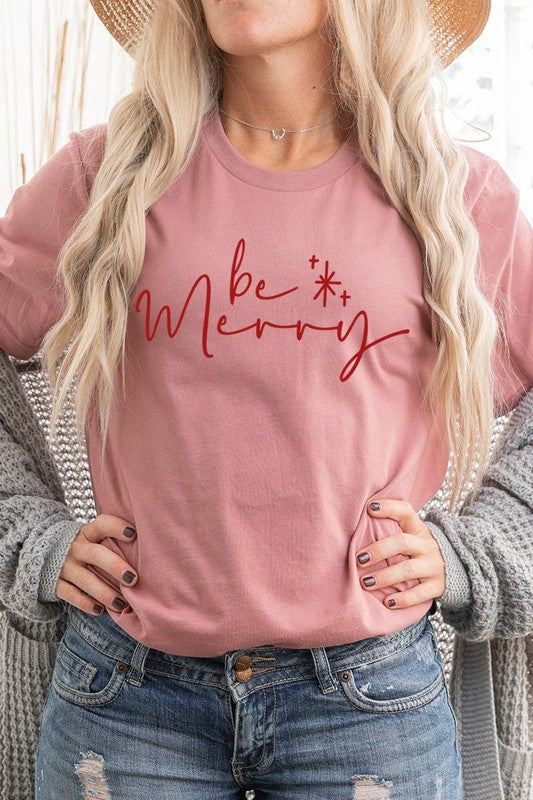 A person wearing the stylish and high-quality Be Merry Graphic Tee, featuring a decorative design with light-colored fabric and bold red text that reads "Be Merry." The unisex crew neck shirt is perfectly paired with them holding a red and black plaid garment.