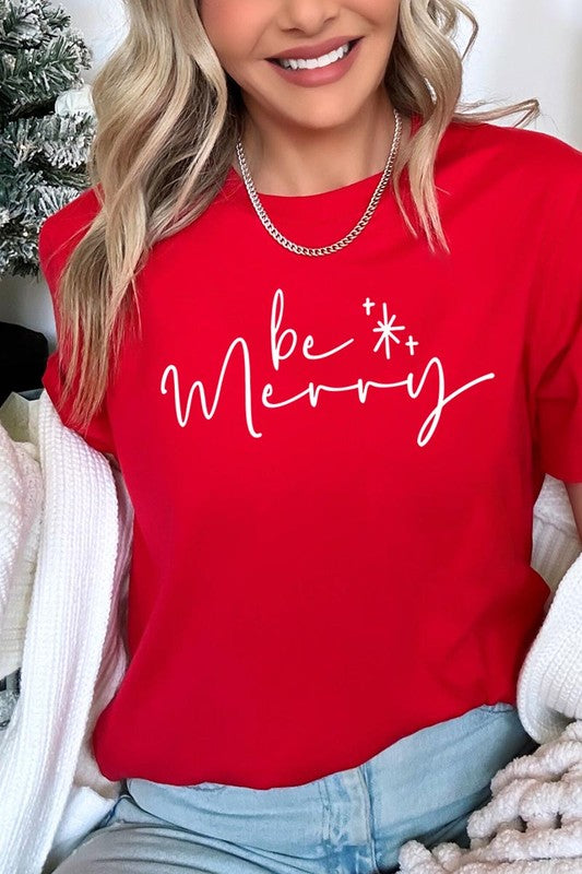 A person wearing the stylish and high-quality Be Merry Graphic Tee, featuring a decorative design with light-colored fabric and bold red text that reads "Be Merry." The unisex crew neck shirt is perfectly paired with them holding a red and black plaid garment.