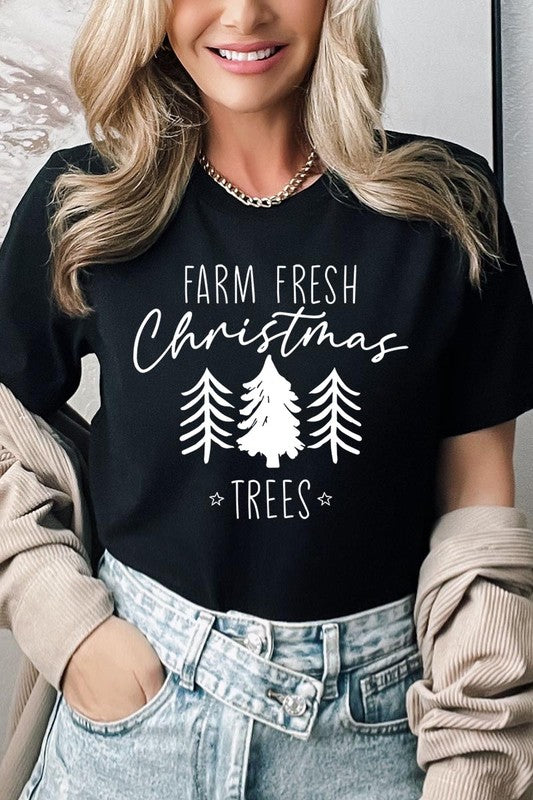 A woman wearing the "Farm Fresh Christmas Trees Graphic Tee" in green, adorned with white text and tree graphics.