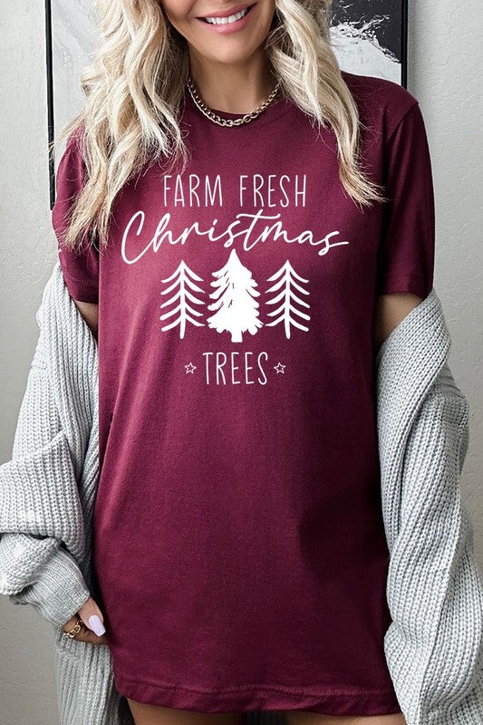 A woman wearing the "Farm Fresh Christmas Trees Graphic Tee" in green, adorned with white text and tree graphics.