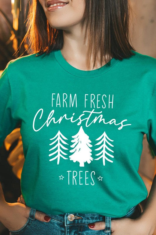 A woman wearing the "Farm Fresh Christmas Trees Graphic Tee" in green, adorned with white text and tree graphics.