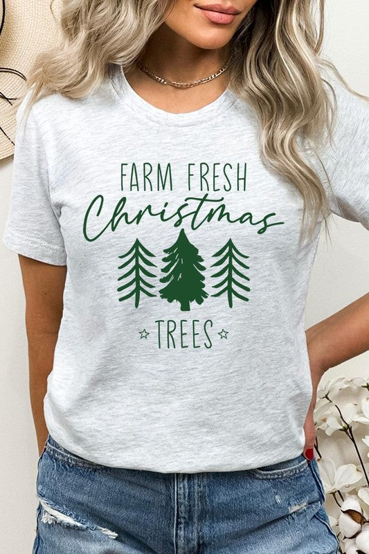 A woman wearing the "Farm Fresh Christmas Trees Graphic Tee" in green, adorned with white text and tree graphics.