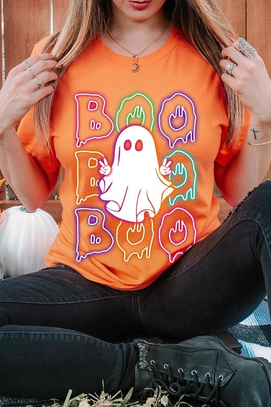 A person in a Boo Neon Halloween Ghost Graphic Tee, featuring neon letters spelling "BOO" with a cartoon ghost in the center. They are wearing light blue jeans and have a beige jacket draped over their arms.