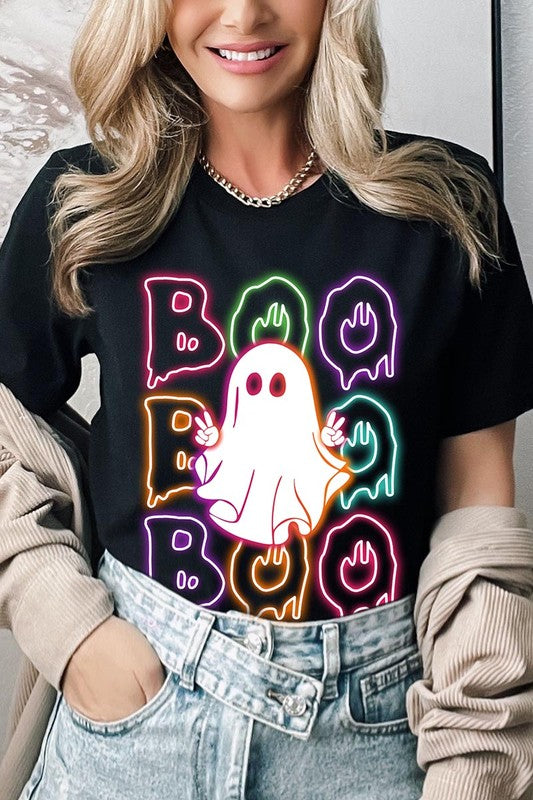 A person in a Boo Neon Halloween Ghost Graphic Tee, featuring neon letters spelling "BOO" with a cartoon ghost in the center. They are wearing light blue jeans and have a beige jacket draped over their arms.