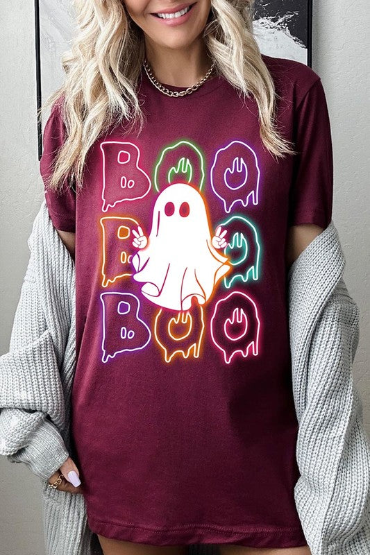 A person in a Boo Neon Halloween Ghost Graphic Tee, featuring neon letters spelling "BOO" with a cartoon ghost in the center. They are wearing light blue jeans and have a beige jacket draped over their arms.