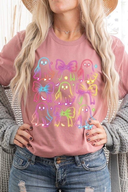 A woman with long, wavy hair and a neutral expression stands against a white brick wall, wearing the Coquette Neon Halloween Ghost Graphic Tee, which features a colorful neon design of ghosts and bows.