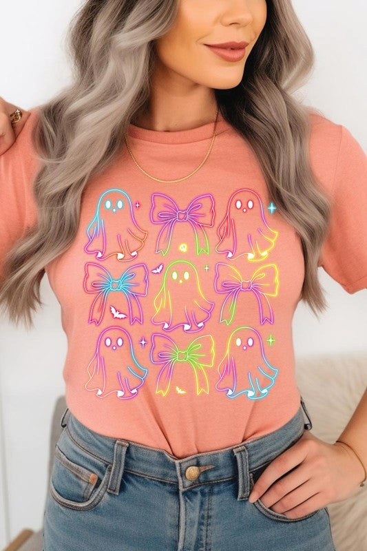 A woman with long, wavy hair and a neutral expression stands against a white brick wall, wearing the Coquette Neon Halloween Ghost Graphic Tee, which features a colorful neon design of ghosts and bows.