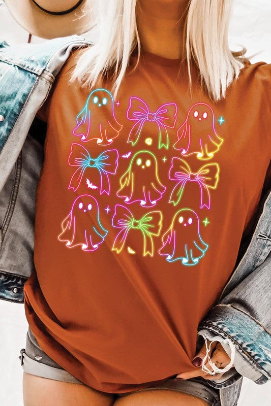 A woman with long, wavy hair and a neutral expression stands against a white brick wall, wearing the Coquette Neon Halloween Ghost Graphic Tee, which features a colorful neon design of ghosts and bows.