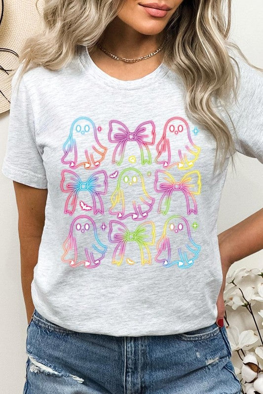 A woman with long, wavy hair and a neutral expression stands against a white brick wall, wearing the Coquette Neon Halloween Ghost Graphic Tee, which features a colorful neon design of ghosts and bows.