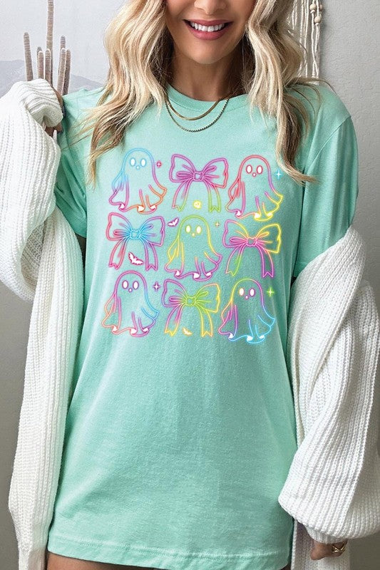 A woman with long, wavy hair and a neutral expression stands against a white brick wall, wearing the Coquette Neon Halloween Ghost Graphic Tee, which features a colorful neon design of ghosts and bows.