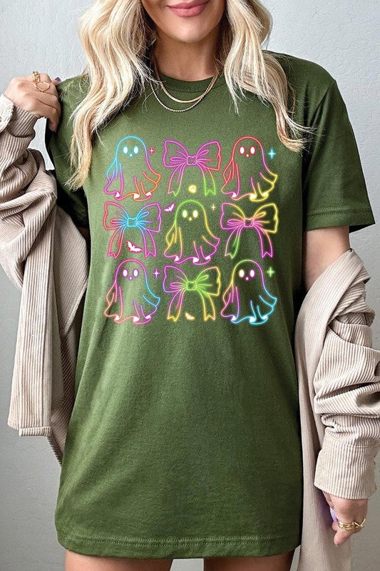 A woman with long, wavy hair and a neutral expression stands against a white brick wall, wearing the Coquette Neon Halloween Ghost Graphic Tee, which features a colorful neon design of ghosts and bows.