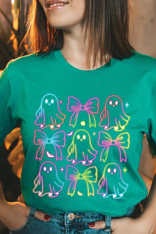 A woman with long, wavy hair and a neutral expression stands against a white brick wall, wearing the Coquette Neon Halloween Ghost Graphic Tee, which features a colorful neon design of ghosts and bows.