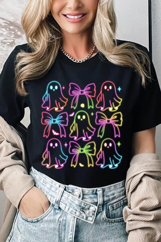 A woman with long, wavy hair and a neutral expression stands against a white brick wall, wearing the Coquette Neon Halloween Ghost Graphic Tee, which features a colorful neon design of ghosts and bows.