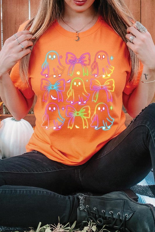 A woman with long, wavy hair and a neutral expression stands against a white brick wall, wearing the Coquette Neon Halloween Ghost Graphic Tee, which features a colorful neon design of ghosts and bows.