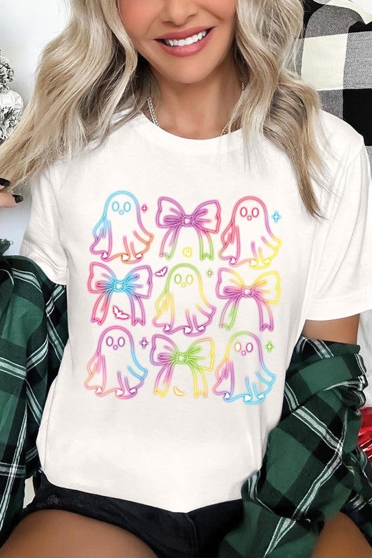 A woman with long, wavy hair and a neutral expression stands against a white brick wall, wearing the Coquette Neon Halloween Ghost Graphic Tee, which features a colorful neon design of ghosts and bows.