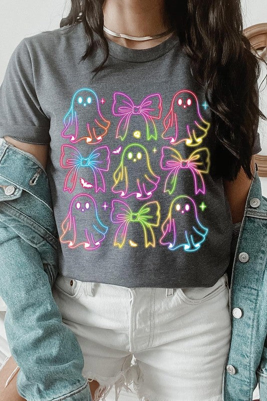 A woman with long, wavy hair and a neutral expression stands against a white brick wall, wearing the Coquette Neon Halloween Ghost Graphic Tee, which features a colorful neon design of ghosts and bows.