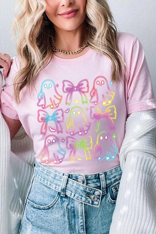 A woman with long, wavy hair and a neutral expression stands against a white brick wall, wearing the Coquette Neon Halloween Ghost Graphic Tee, which features a colorful neon design of ghosts and bows.