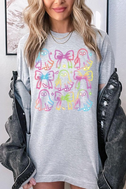 A woman with long, wavy hair and a neutral expression stands against a white brick wall, wearing the Coquette Neon Halloween Ghost Graphic Tee, which features a colorful neon design of ghosts and bows.