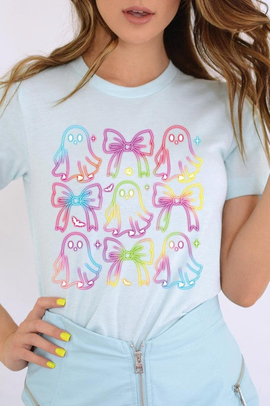 A woman with long, wavy hair and a neutral expression stands against a white brick wall, wearing the Coquette Neon Halloween Ghost Graphic Tee, which features a colorful neon design of ghosts and bows.