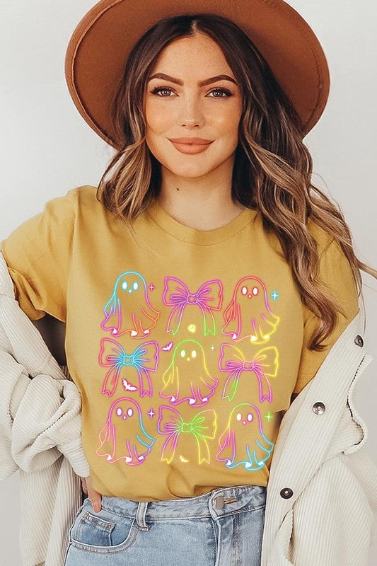 A woman with long, wavy hair and a neutral expression stands against a white brick wall, wearing the Coquette Neon Halloween Ghost Graphic Tee, which features a colorful neon design of ghosts and bows.