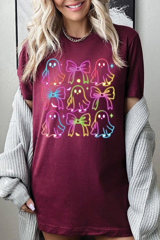 A woman with long, wavy hair and a neutral expression stands against a white brick wall, wearing the Coquette Neon Halloween Ghost Graphic Tee, which features a colorful neon design of ghosts and bows.