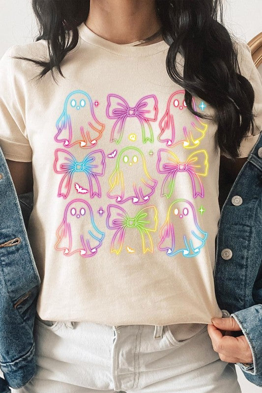 A woman with long, wavy hair and a neutral expression stands against a white brick wall, wearing the Coquette Neon Halloween Ghost Graphic Tee, which features a colorful neon design of ghosts and bows.