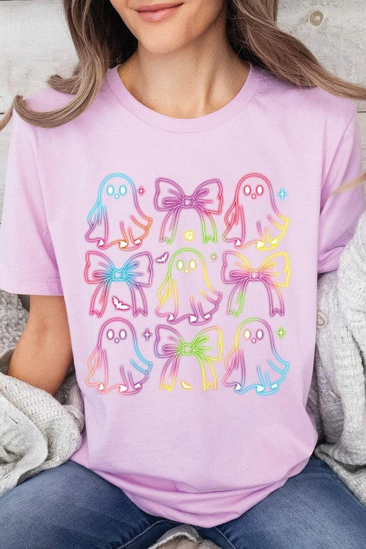 A woman with long, wavy hair and a neutral expression stands against a white brick wall, wearing the Coquette Neon Halloween Ghost Graphic Tee, which features a colorful neon design of ghosts and bows.