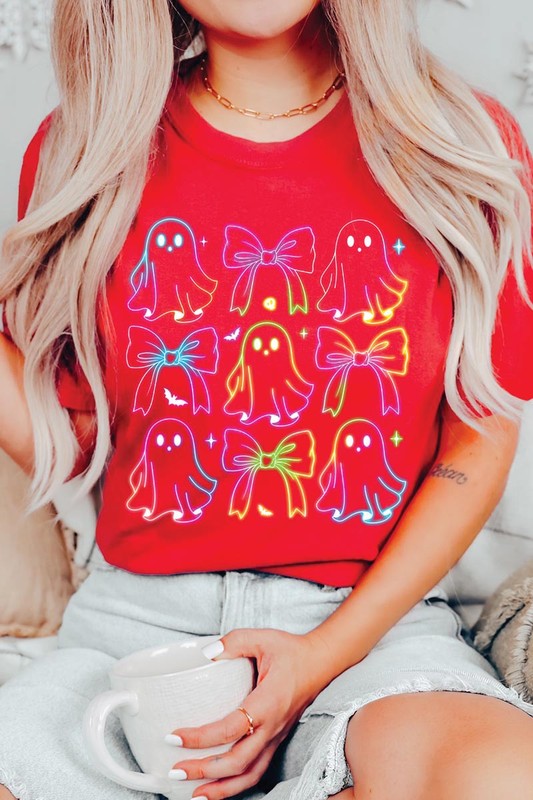 A woman with long, wavy hair and a neutral expression stands against a white brick wall, wearing the Coquette Neon Halloween Ghost Graphic Tee, which features a colorful neon design of ghosts and bows.