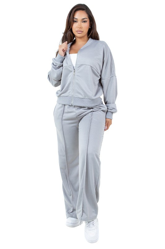 A person stands in a relaxed pose, wearing the WOMEN FASHION SWEATSUIT TWO PIECE PANT SET, which features a green zip-up jacket with a front-zip closure and matching green pants with an elasticized waist over a white top, paired with white sneakers.