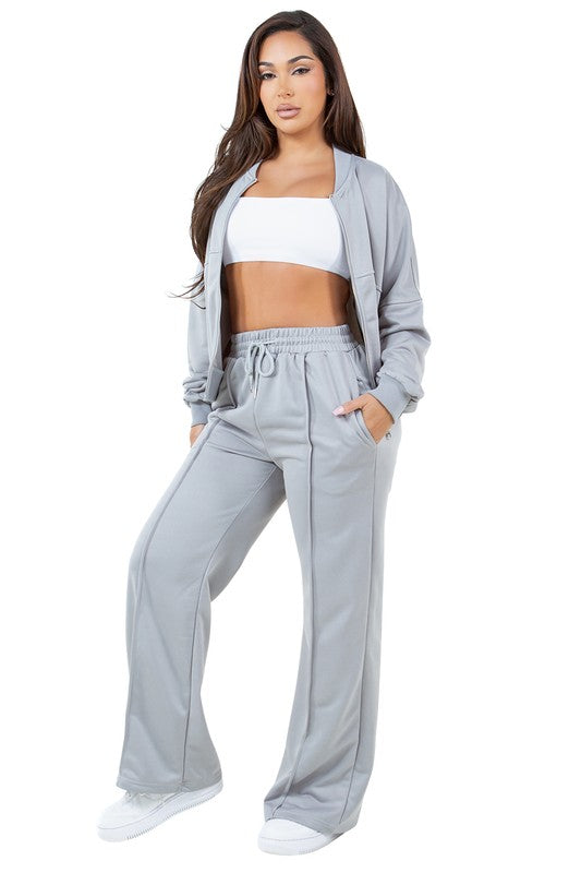 A person stands in a relaxed pose, wearing the WOMEN FASHION SWEATSUIT TWO PIECE PANT SET, which features a green zip-up jacket with a front-zip closure and matching green pants with an elasticized waist over a white top, paired with white sneakers.