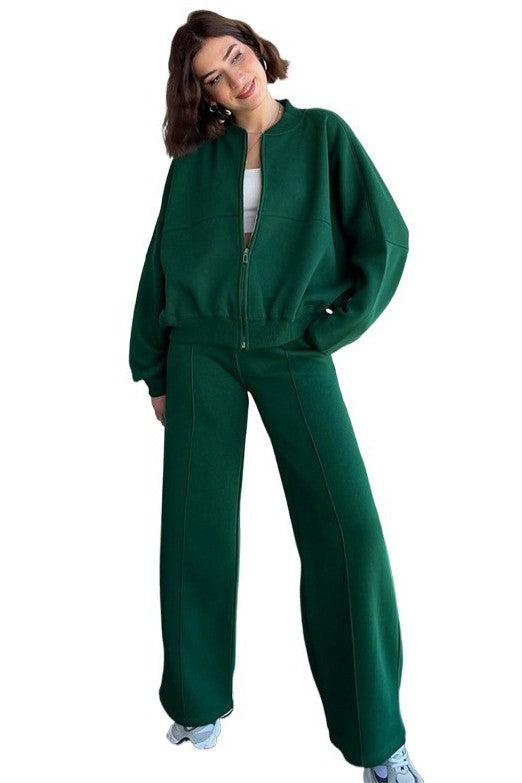 A person stands in a relaxed pose, wearing the WOMEN FASHION SWEATSUIT TWO PIECE PANT SET, which features a green zip-up jacket with a front-zip closure and matching green pants with an elasticized waist over a white top, paired with white sneakers.