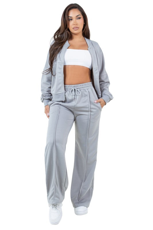 A person stands in a relaxed pose, wearing the WOMEN FASHION SWEATSUIT TWO PIECE PANT SET, which features a green zip-up jacket with a front-zip closure and matching green pants with an elasticized waist over a white top, paired with white sneakers.