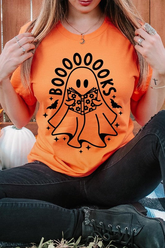 A person in a Cute Ghost Book Lover Graphic Tee featuring the text "BOOOOOOKS" holds a "Spells & Potions" book, set against a background of small bat decorations.