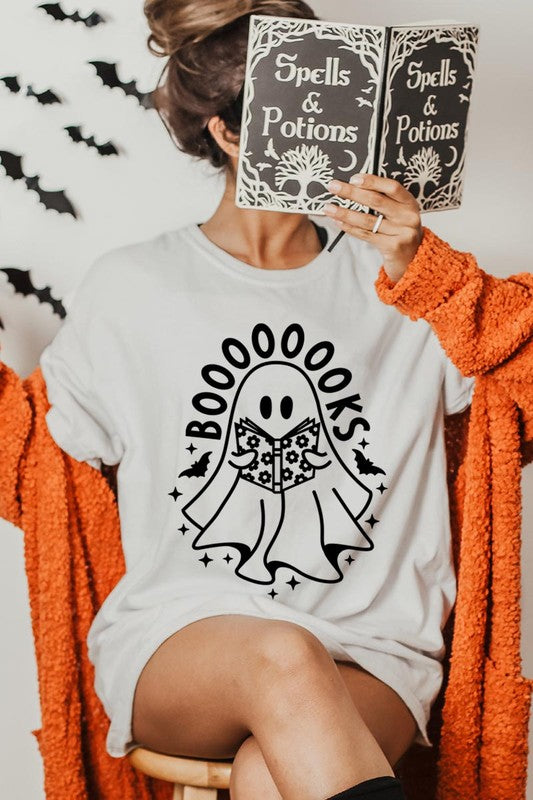 A person in a Cute Ghost Book Lover Graphic Tee featuring the text "BOOOOOOKS" holds a "Spells & Potions" book, set against a background of small bat decorations.