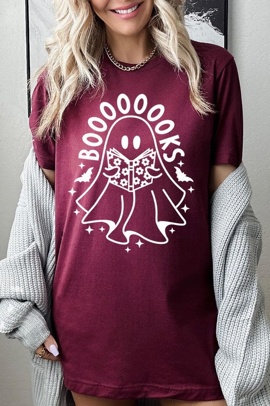 A person in a Cute Ghost Book Lover Graphic Tee featuring the text "BOOOOOOKS" holds a "Spells & Potions" book, set against a background of small bat decorations.