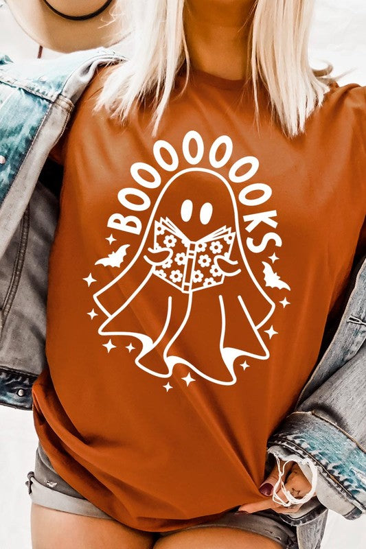 A person in a Cute Ghost Book Lover Graphic Tee featuring the text "BOOOOOOKS" holds a "Spells & Potions" book, set against a background of small bat decorations.