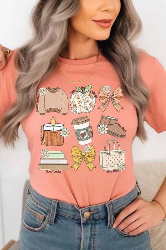 A person in a grey **Fall Girly Doodle Graphic Tee**, featuring charming illustrations of a sweater, apple, books, bows, coffee cup, candle, and tote bag, holds an orange mug while wearing a red cardigan and ripped jeans.