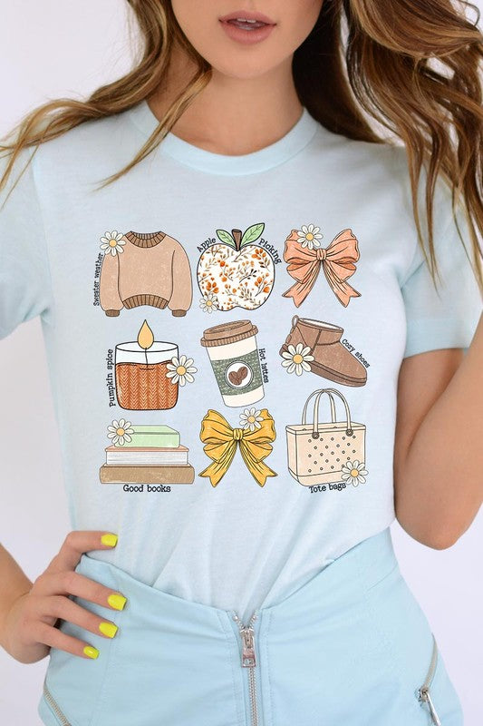 A person in a grey **Fall Girly Doodle Graphic Tee**, featuring charming illustrations of a sweater, apple, books, bows, coffee cup, candle, and tote bag, holds an orange mug while wearing a red cardigan and ripped jeans.