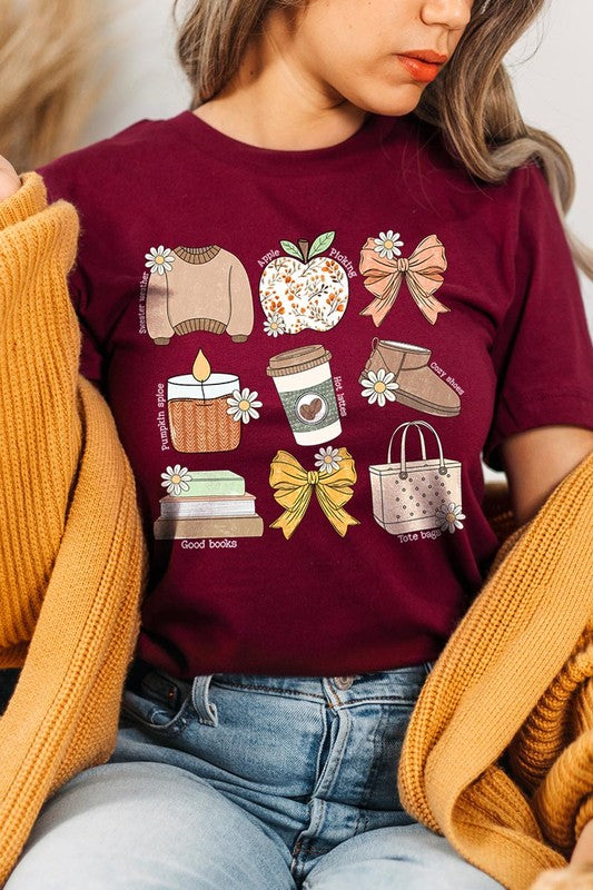 A person in a grey **Fall Girly Doodle Graphic Tee**, featuring charming illustrations of a sweater, apple, books, bows, coffee cup, candle, and tote bag, holds an orange mug while wearing a red cardigan and ripped jeans.