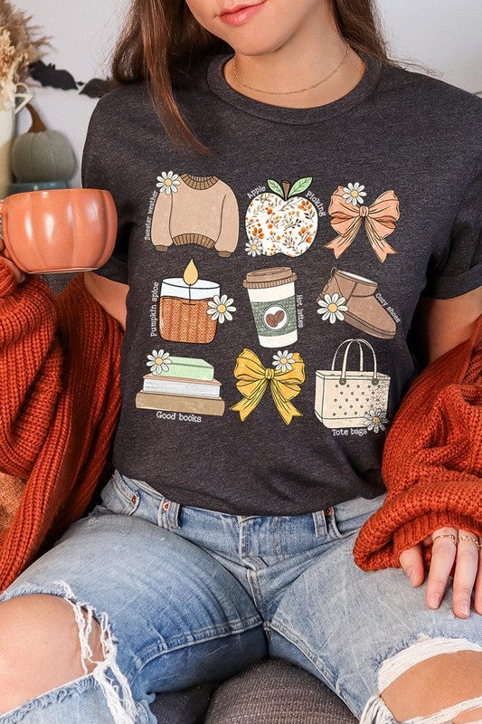 A person in a grey **Fall Girly Doodle Graphic Tee**, featuring charming illustrations of a sweater, apple, books, bows, coffee cup, candle, and tote bag, holds an orange mug while wearing a red cardigan and ripped jeans.