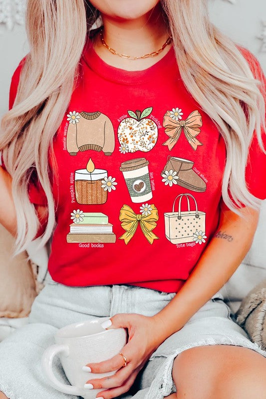 A person in a grey **Fall Girly Doodle Graphic Tee**, featuring charming illustrations of a sweater, apple, books, bows, coffee cup, candle, and tote bag, holds an orange mug while wearing a red cardigan and ripped jeans.