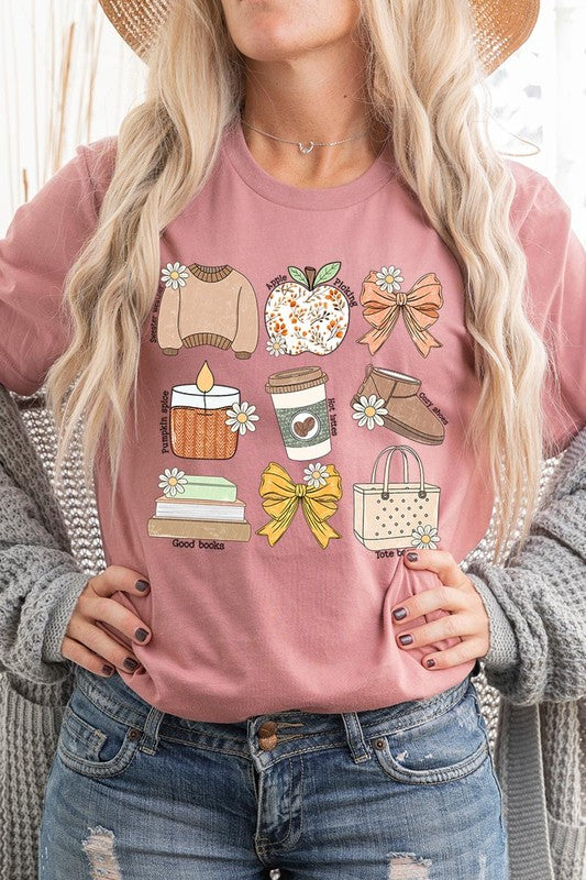 A person in a grey **Fall Girly Doodle Graphic Tee**, featuring charming illustrations of a sweater, apple, books, bows, coffee cup, candle, and tote bag, holds an orange mug while wearing a red cardigan and ripped jeans.