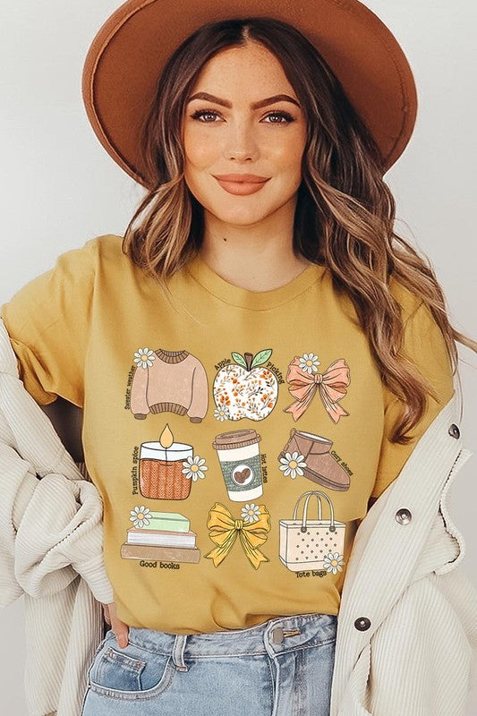 A person in a grey **Fall Girly Doodle Graphic Tee**, featuring charming illustrations of a sweater, apple, books, bows, coffee cup, candle, and tote bag, holds an orange mug while wearing a red cardigan and ripped jeans.