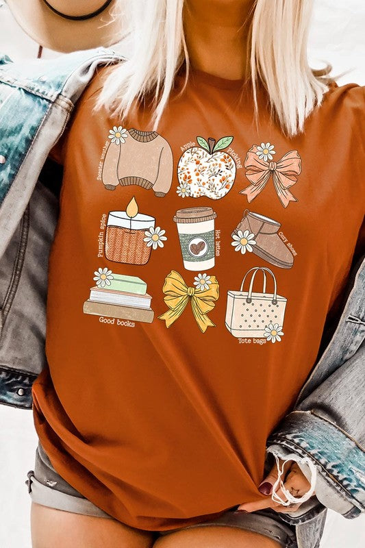 A person in a grey **Fall Girly Doodle Graphic Tee**, featuring charming illustrations of a sweater, apple, books, bows, coffee cup, candle, and tote bag, holds an orange mug while wearing a red cardigan and ripped jeans.