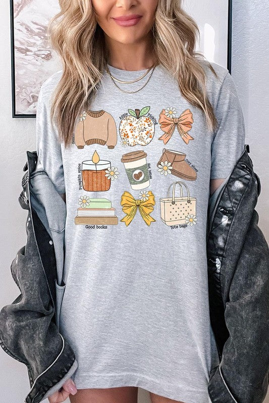 A person in a grey **Fall Girly Doodle Graphic Tee**, featuring charming illustrations of a sweater, apple, books, bows, coffee cup, candle, and tote bag, holds an orange mug while wearing a red cardigan and ripped jeans.