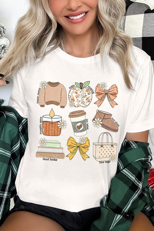 A person in a grey **Fall Girly Doodle Graphic Tee**, featuring charming illustrations of a sweater, apple, books, bows, coffee cup, candle, and tote bag, holds an orange mug while wearing a red cardigan and ripped jeans.