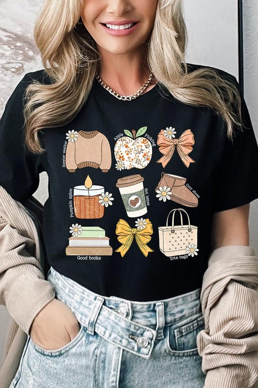 A person in a grey **Fall Girly Doodle Graphic Tee**, featuring charming illustrations of a sweater, apple, books, bows, coffee cup, candle, and tote bag, holds an orange mug while wearing a red cardigan and ripped jeans.