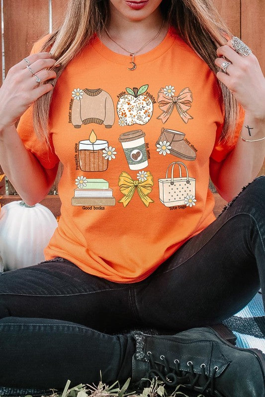 A person in a grey **Fall Girly Doodle Graphic Tee**, featuring charming illustrations of a sweater, apple, books, bows, coffee cup, candle, and tote bag, holds an orange mug while wearing a red cardigan and ripped jeans.