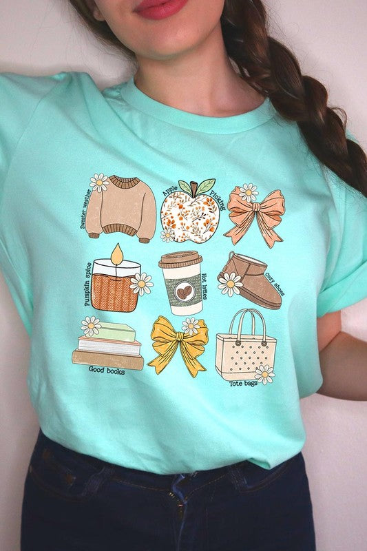 A person in a grey **Fall Girly Doodle Graphic Tee**, featuring charming illustrations of a sweater, apple, books, bows, coffee cup, candle, and tote bag, holds an orange mug while wearing a red cardigan and ripped jeans.