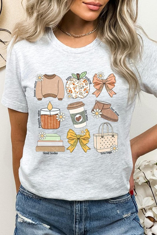 A person in a grey **Fall Girly Doodle Graphic Tee**, featuring charming illustrations of a sweater, apple, books, bows, coffee cup, candle, and tote bag, holds an orange mug while wearing a red cardigan and ripped jeans.